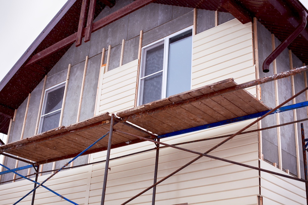 Scaffolding around house with beige siding covering walls. Construction site.
